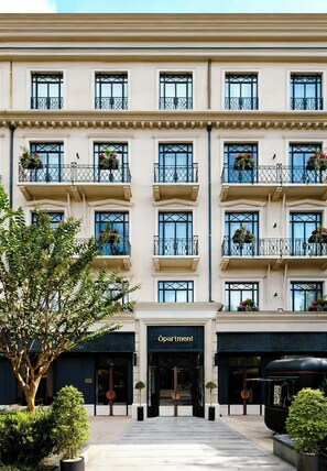 Un hotel de varios pisos con una fachada clásica, múltiples balcones con macetas de flores y una entrada destacada con un cartel que dice "Apartment".