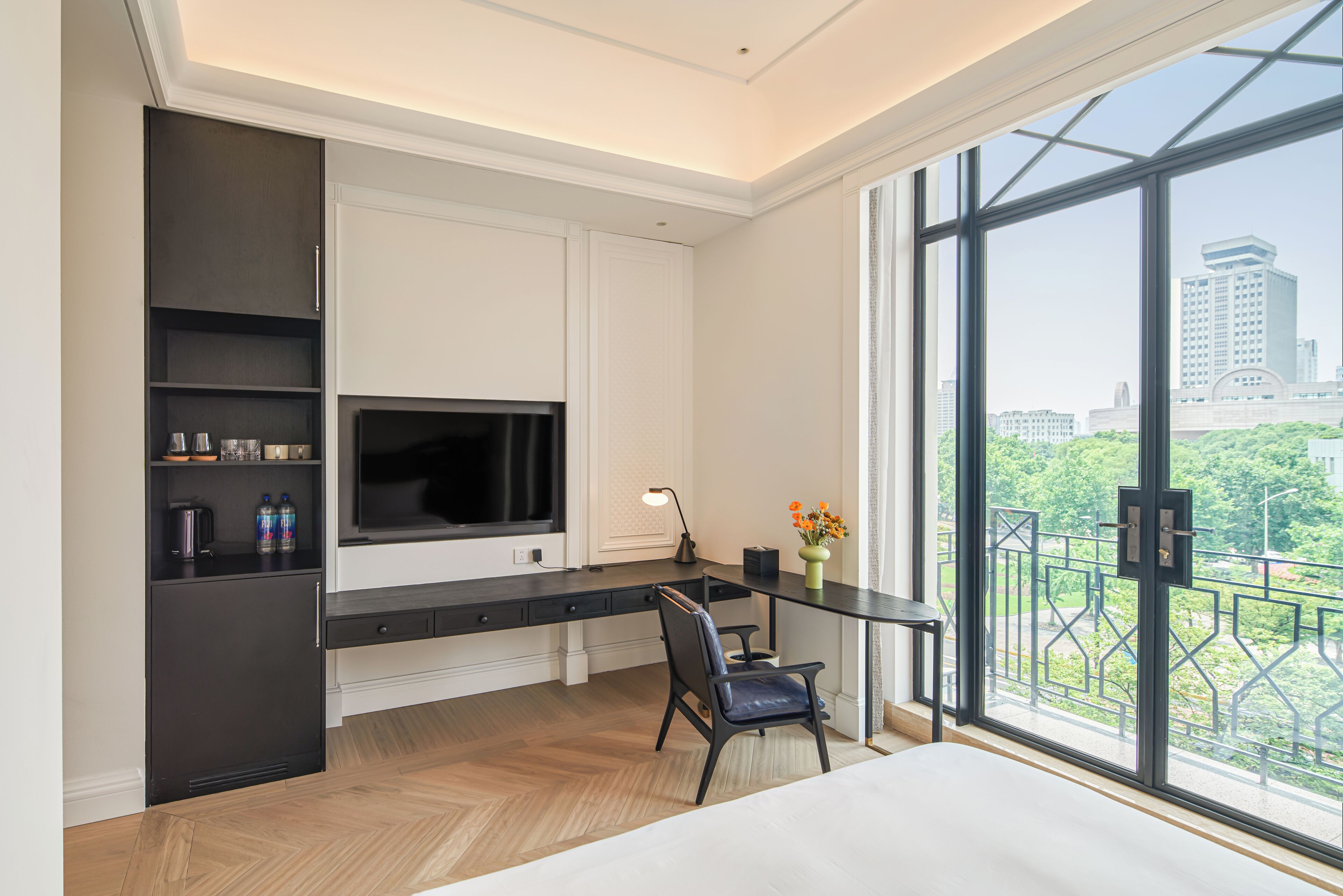 A modern hotel room with a large window, a desk with a flat-screen TV, a chair, and a minibar.