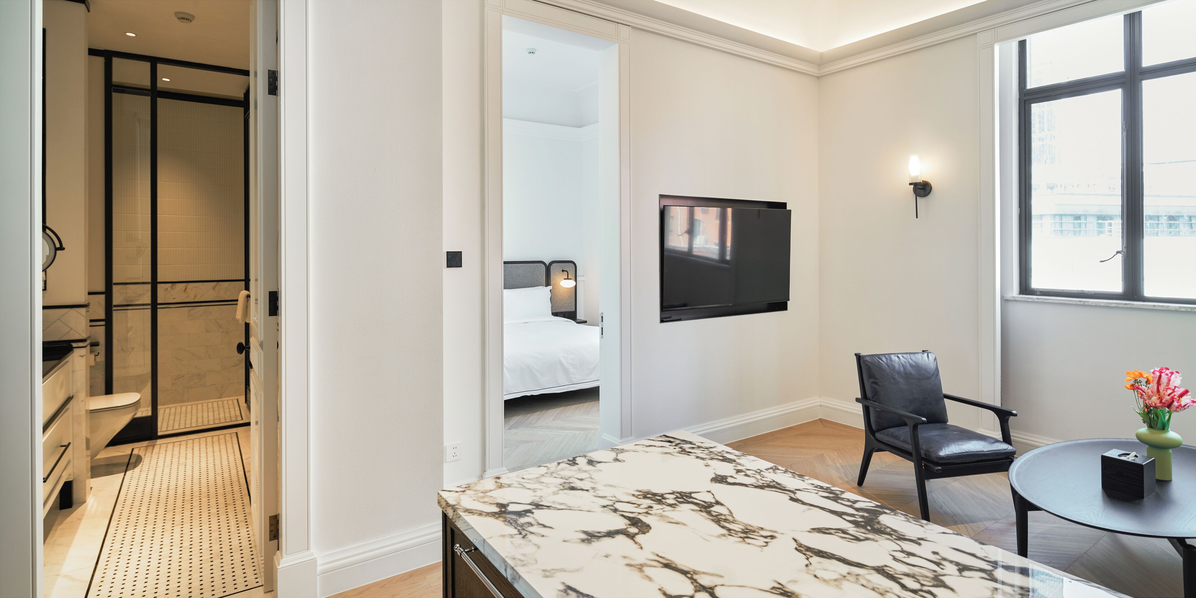 A modern hotel room with a marble-topped desk, a black leather chair, a bed with white bedding, a flat-screen TV mounted on the wall, and a large window offering a view of the city.