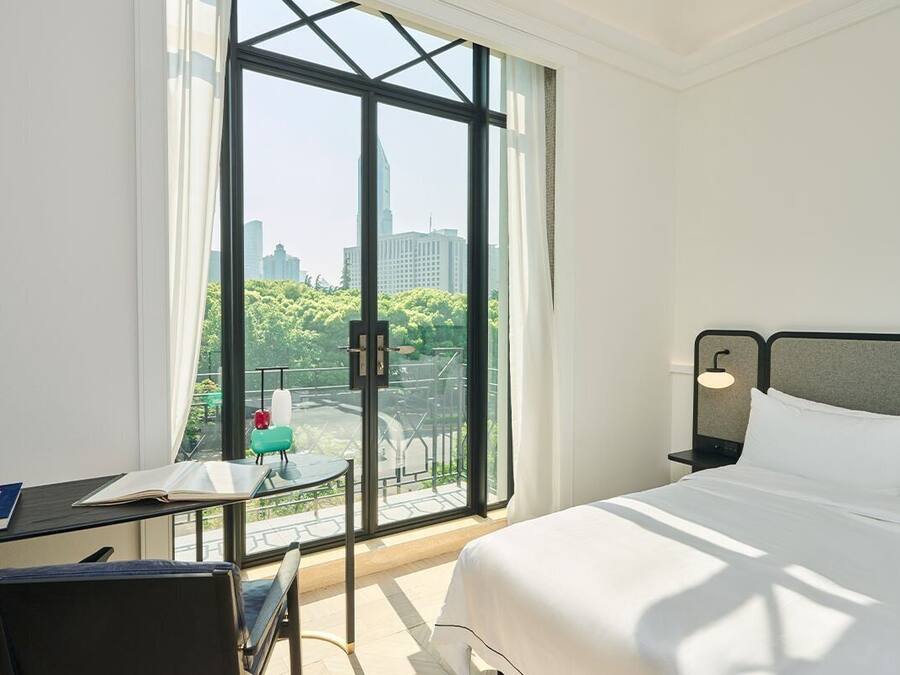 A hotel room with a large window, a bed, a desk with a chair, and a small table with a lamp.