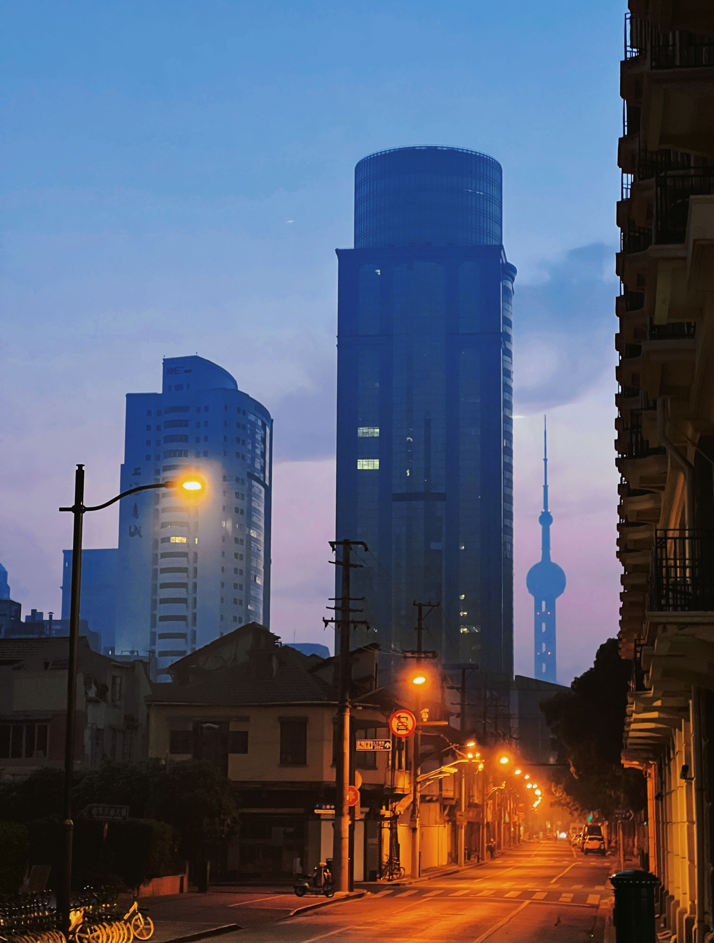 A cityscape with modern buildings, a prominent tower, and a street with streetlights.