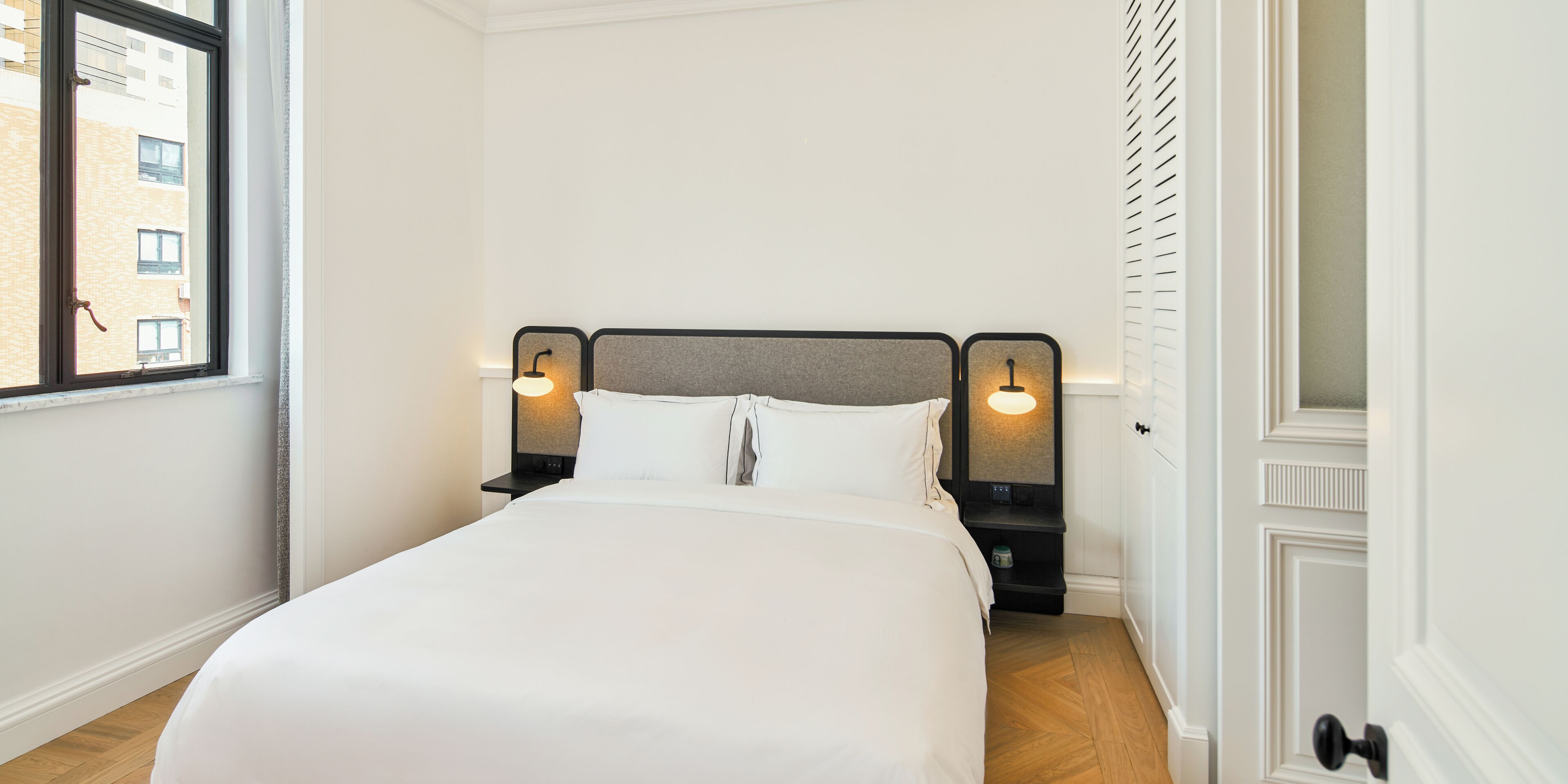 Una cama bien tendida en una habitación de hotel, con una ventana, dos mesitas de noche con lámparas y una puerta con manija.