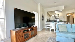 Un salon doté d’un grand téléviseur à écran plat, d’un meuble de rangement en bois, d’un canapé bleu et d’un coin repas avec des chaises blanches.
