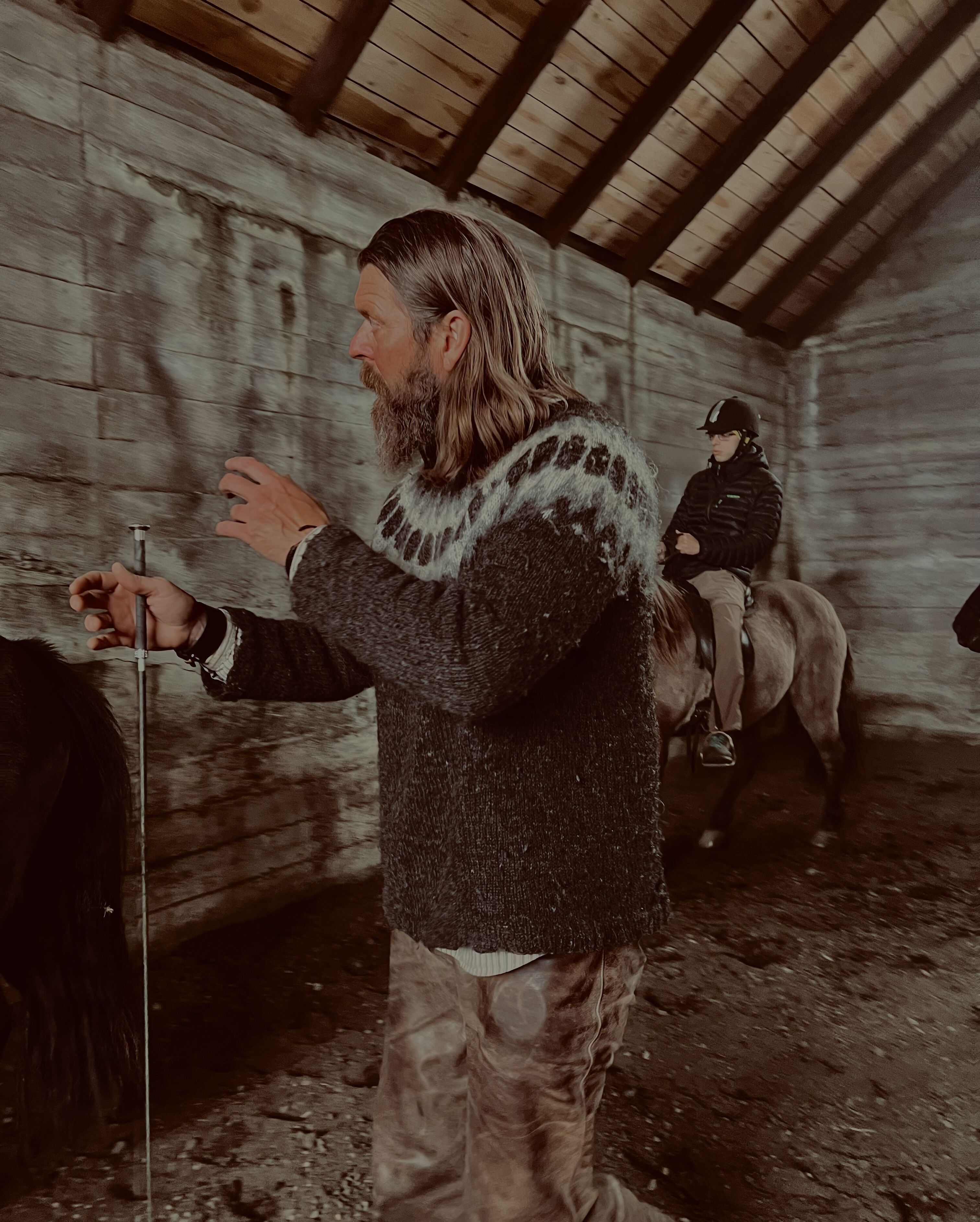 A person in a stable with a horse, wearing a sweater and holding a stick.