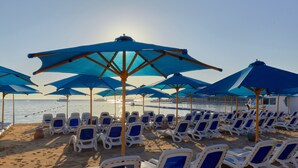 A beach with rows of white lounge chairs and blue umbrellas, arranged for sunbathing. - Naama Bay Suites & Spa (Sharm El Sheikh)