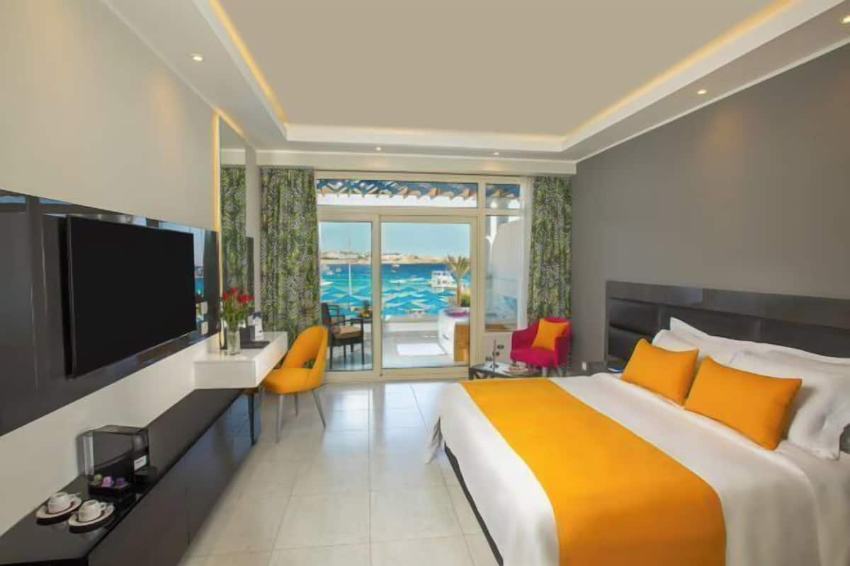 A hotel room with a large bed, a TV, a desk, and a view of the sea.