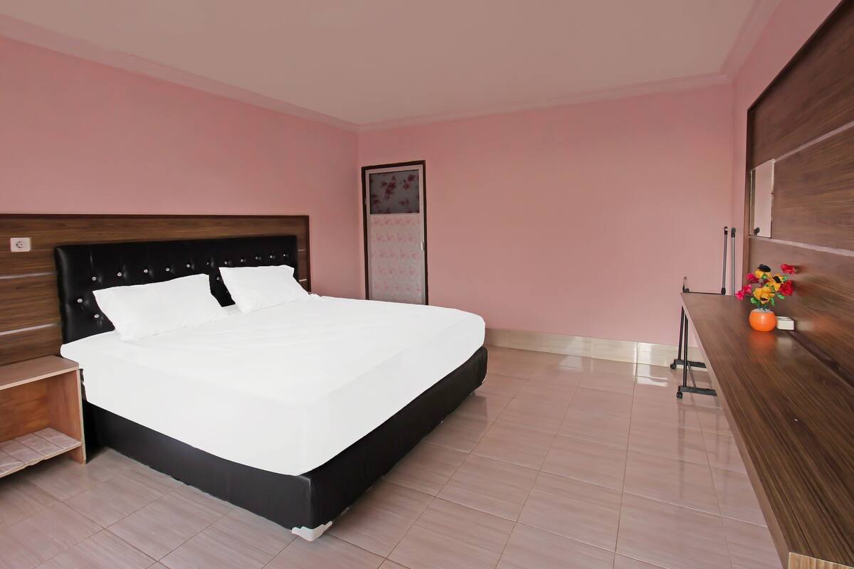 Ein ordentlich eingerichtetes Schlafzimmer mit einem großen Bett, einem Nachttisch aus Holz, einem Schreibtisch mit Blumen und einem gerahmten Bild an der Wand.