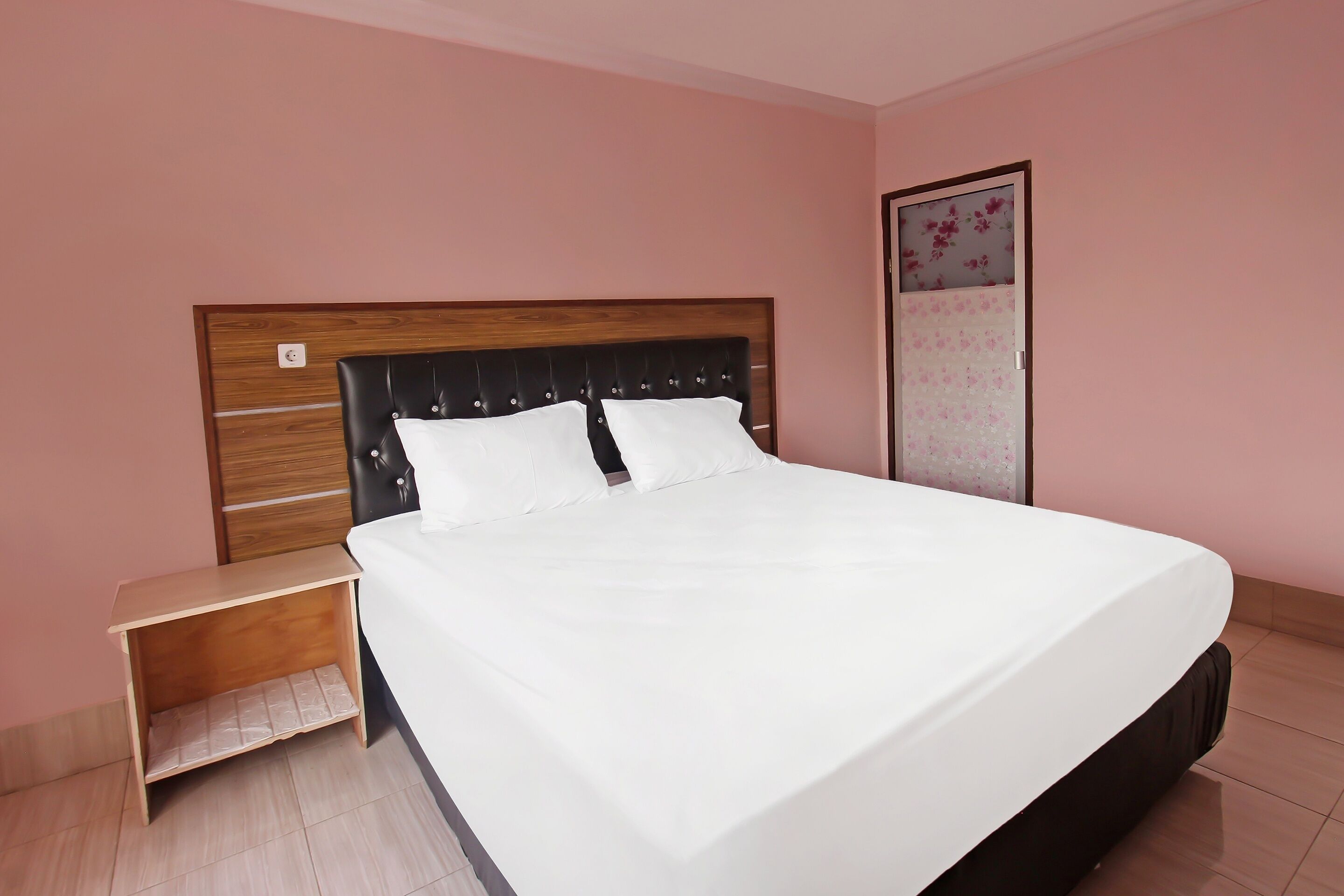 Una cama bien hecha con cabecera de madera y ropa de cama blanca en una habitación con paredes rosadas y una mesita de noche de madera.