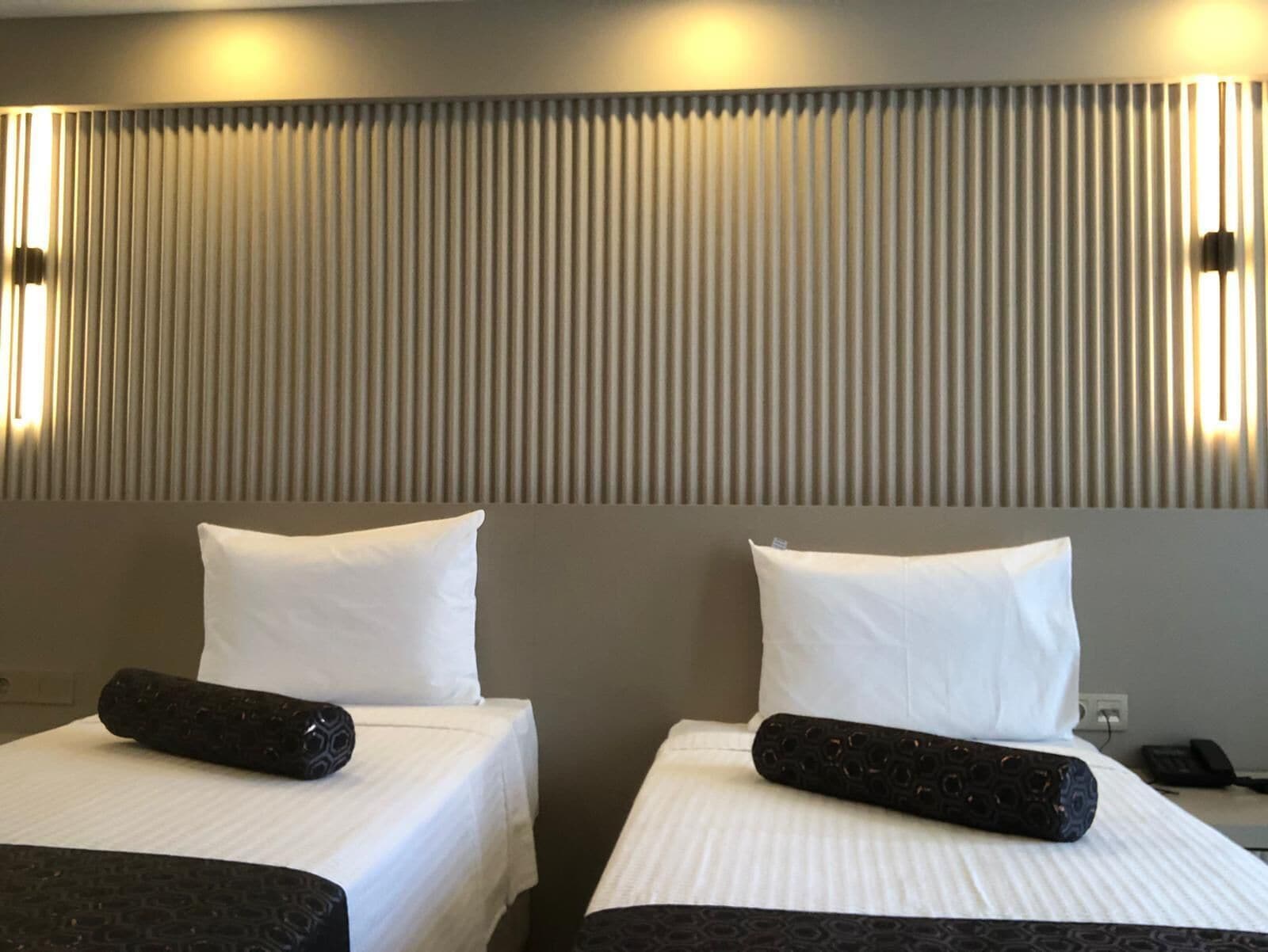 Une chambre d’hôtel avec deux lits, chacun agrémenté d’un oreiller blanc et d’un objet cylindrique noir posé sur le lit.