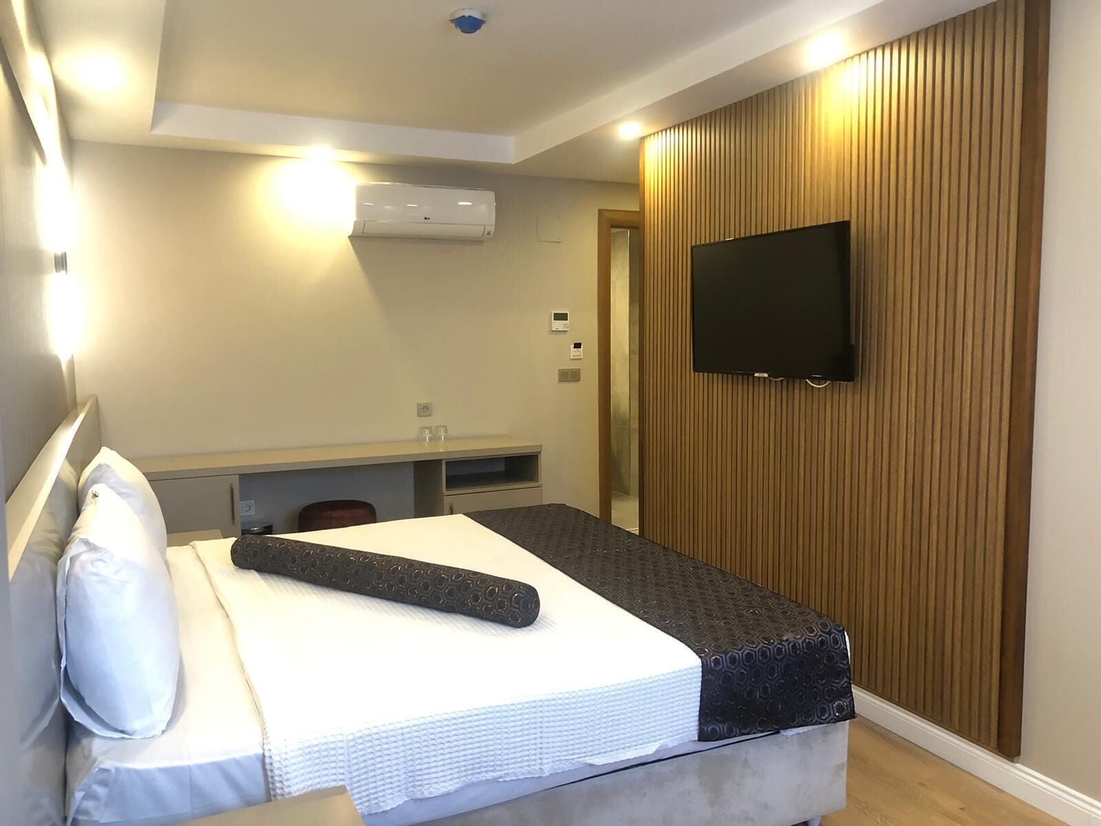 Habitación de hotel con cama, televisor de pantalla plana montado en la pared, panel de pared de madera y unidad de aire acondicionado instalada en el techo.