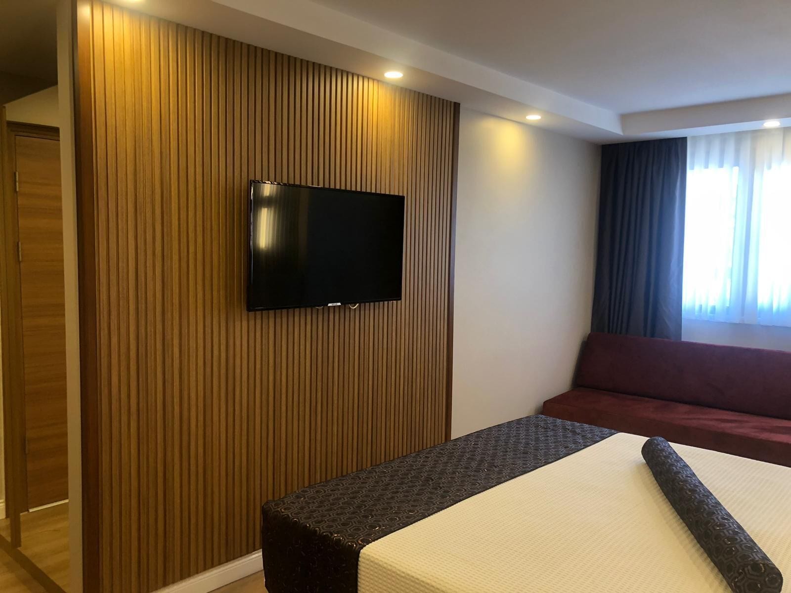 A hotel room with a flat-screen TV mounted on a wooden panel wall, a bed with a white mattress and a dark headboard, and a red sofa.