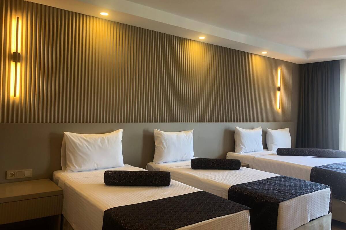 Habitación de hotel con tres camas, cada una con ropa blanca y cabeceras negras. La habitación presenta un diseño moderno con listones de madera verticales en la pared y luces empotradas en el techo.