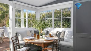 A sunlit dining area with a wooden table set for four, surrounded by white cushioned seating. - Werona Cottage – enchanting home (Burrawang)