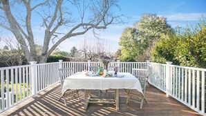 Une terrasse en bois avec une table blanche dressée pour un repas, entourée de rampes blanches et d’arbres.