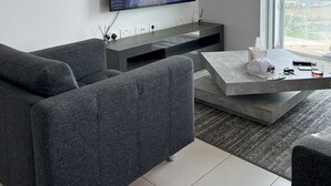 Ein modernes Wohnzimmer mit grauem Sofa, einem Flachbildfernseher an der Wand, einem Couchtisch und einem großen Fenster mit Blick auf die Stadt.