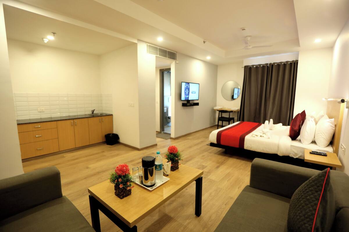 Une chambre d’hôtel moderne avec un plancher en bois, un lit recouvert d’une couverture rouge, un canapé, une petite table sur laquelle se trouve un plateau avec divers objets, et un téléviseur fixé au mur.