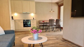 A modern living room with a sofa, a round table with a vase of flowers, a dining area with chairs, and a kitchen with cabinets and a stove.