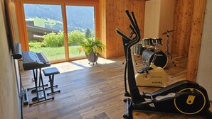 A room with a piano, exercise equipment, a drum set, and a view of a green landscape through large windows.