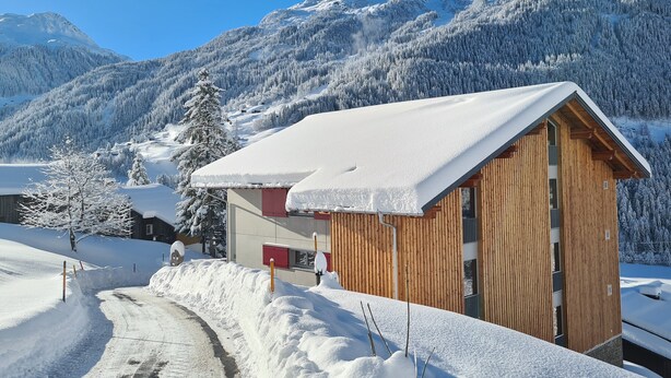 A wooden cabin with a sloped roof, surrounded by snow-covered trees and mountains in the background.