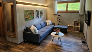 Una sala de estar moderna con un sofá gris, una mesa de centro redonda y piso de madera. Hay tres cuadros enmarcados en la pared, una ventana con cortinas y un pequeño estante con objetos decorativos.