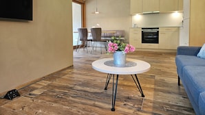Una sala de estar moderna con un televisor de pantalla plana montado en la pared, una mesa de centro redonda con base metálica, un sofá azul y una zona de comedor con una mesa y sillas.
