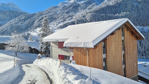 Una cabaña de madera con techo inclinado, rodeada de árboles nevados y montañas al fondo.