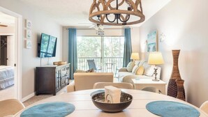 Habitación de hotel con mesa de comedor, sillas, un sofá, televisor, balcón con cortinas y una lámpara decorativa.