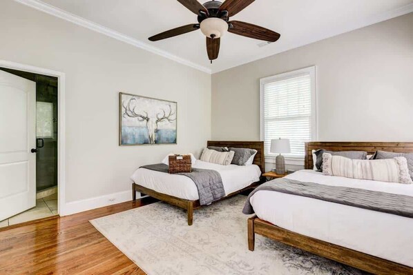 A bedroom with two beds, a ceiling fan, a painting, and a window with blinds.