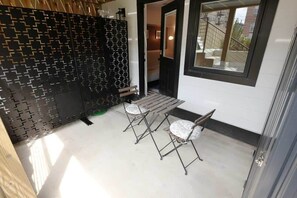A small outdoor seating area with two chairs and a table, a black patterned wall, and a view of the street through a window.