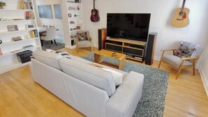 A living room with a white sofa, a TV, a guitar, and a bookshelf. - Meridian Row | Sleeps 7 | Off St Parking! (Washington)