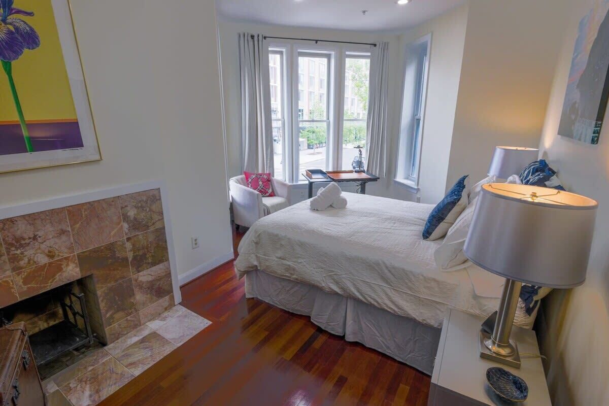 A bedroom with a fireplace, a bed with white bedding, a nightstand with a lamp, and a window with curtains.
