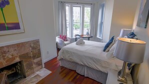 A bedroom with a fireplace, a bed with white bedding, a nightstand with a lamp, and a window with curtains.