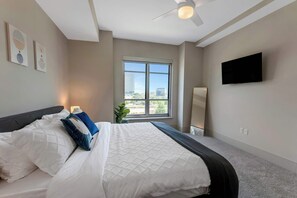 A hotel room with a bed, a ceiling fan, a window with a city view, a flat-screen TV, and artwork on the wall.