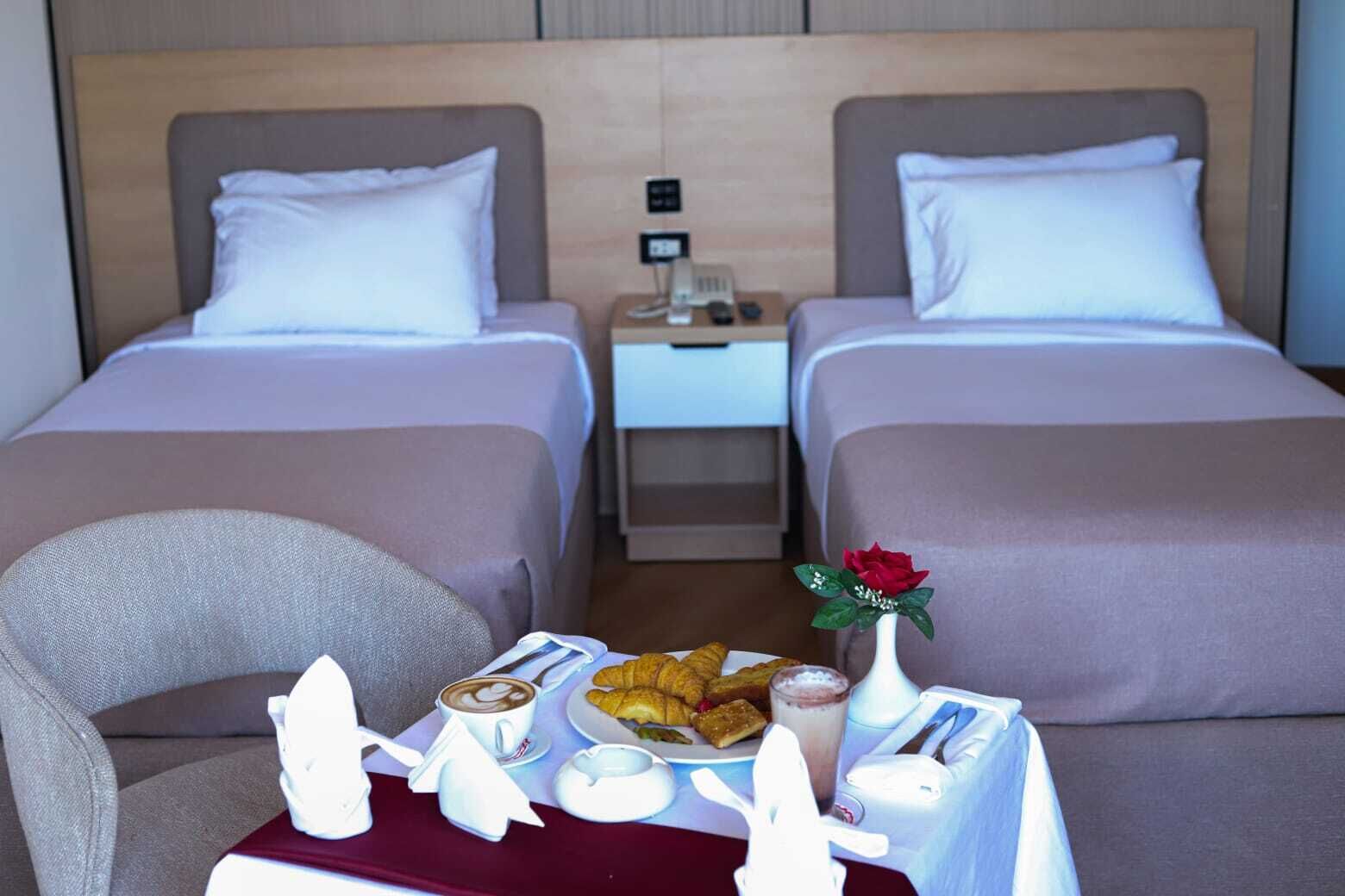 Et hotelværelse med to senge, et natbord med telefon og lampe, samt et morgenmadsserveringsfad med croissanter, kaffe og en lille vase med en blomst.