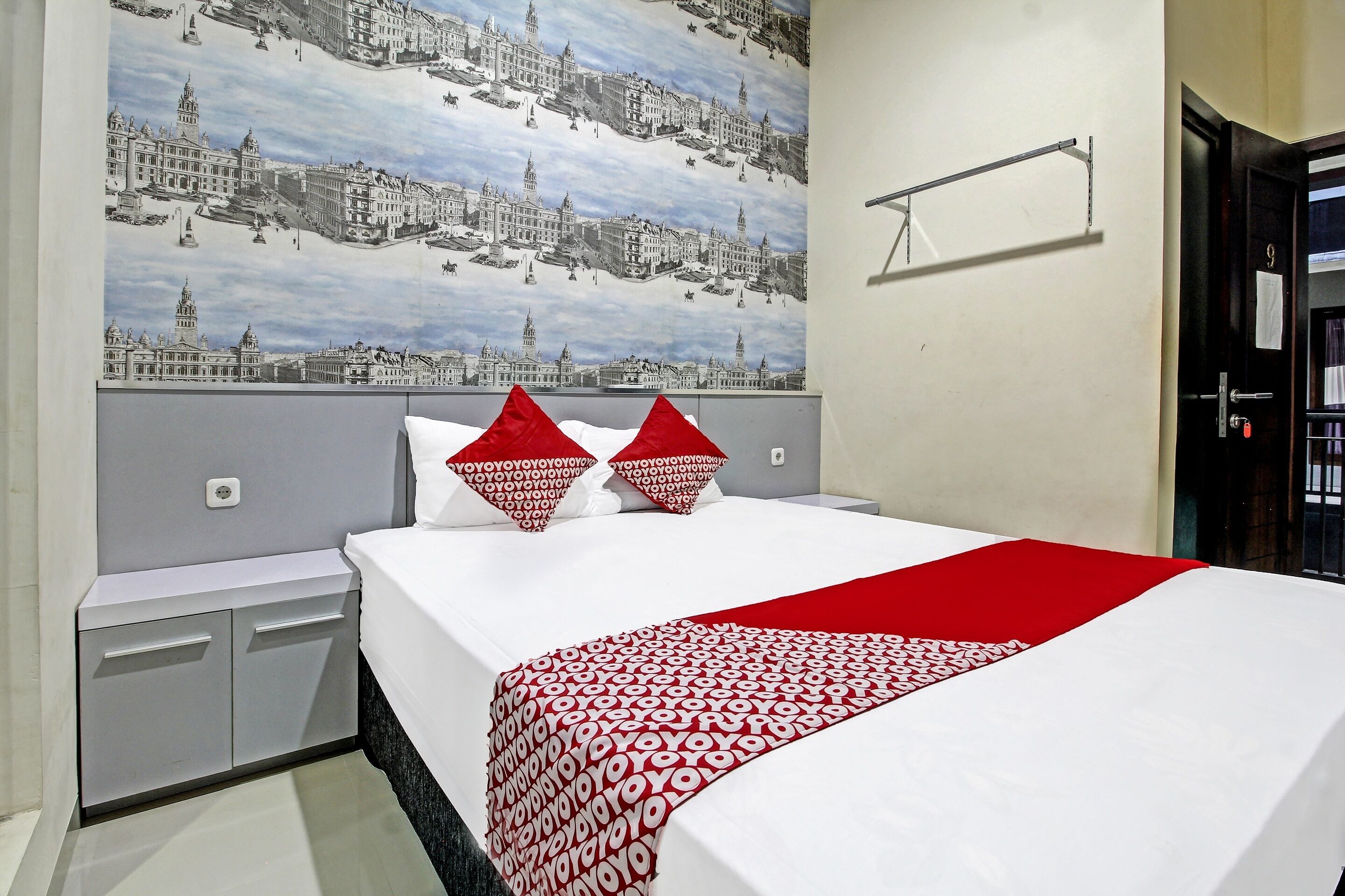 Quarto de hotel com cama, mesa de cabeceira, prateleira na parede e porta.