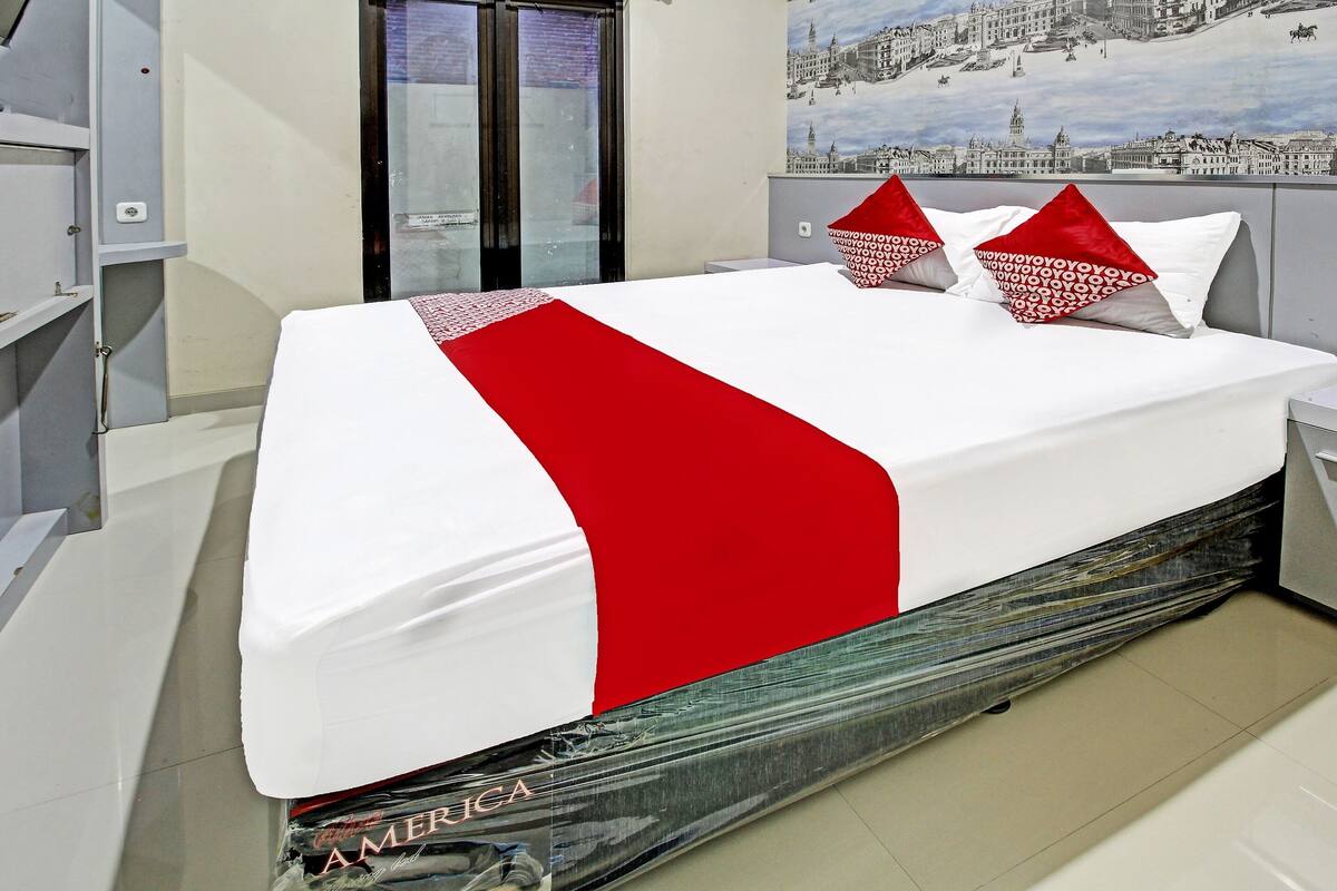 Habitación de hotel con cama, mesita de noche, espejo y ventana con vista panorámica de la ciudad.