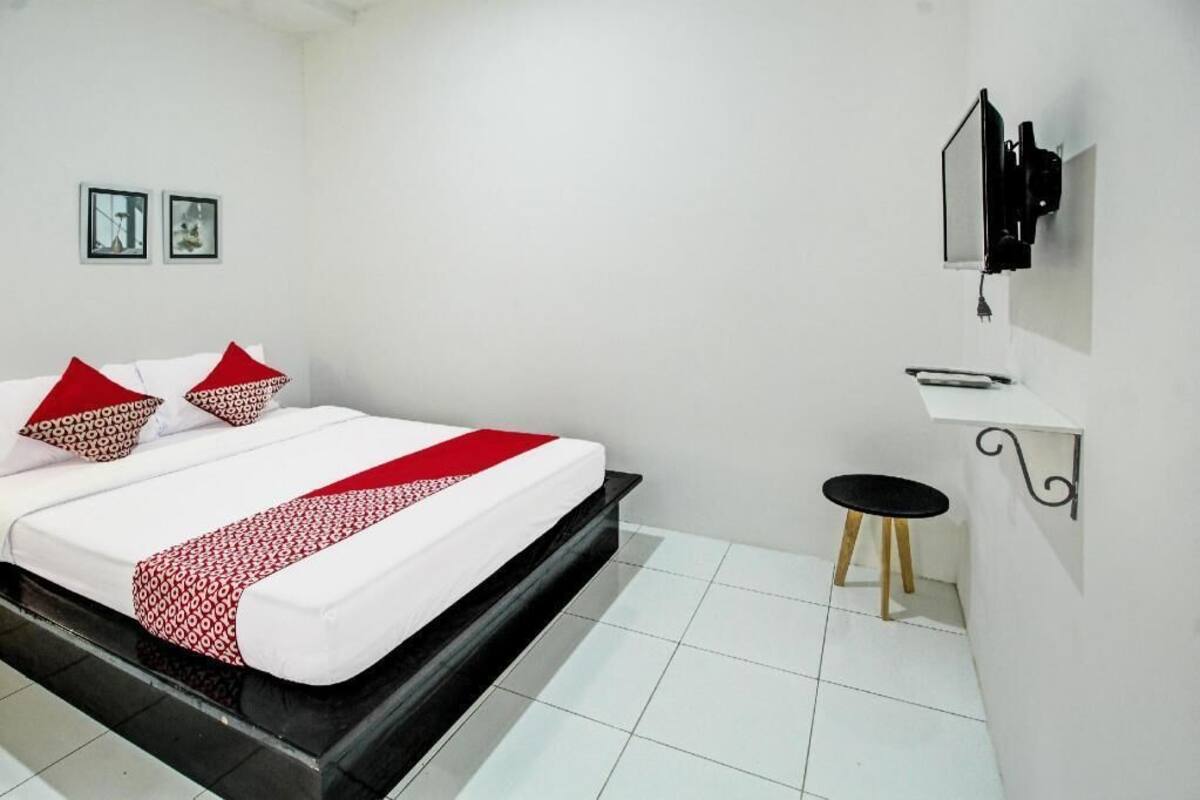 Habitación de hotel con una cama, una mesita pequeña, un televisor montado en la pared y dos cuadros enmarcados.