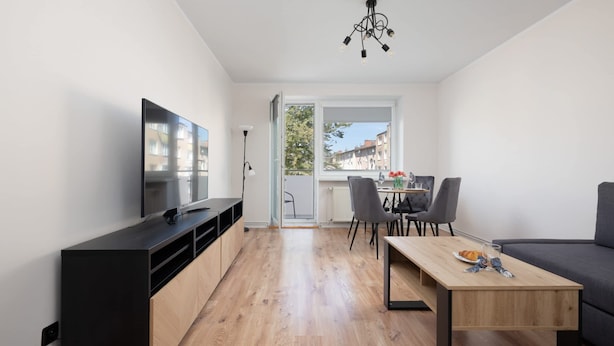 A modern living room with a TV, wooden flooring, a dining table with chairs, a sofa, and a view through a large window.