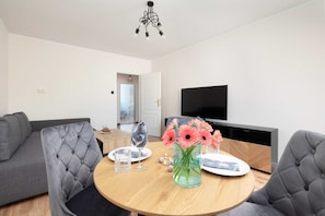 A modern living room with a round wooden table, grey upholstered chairs, a flat-screen TV, and a decorative vase with flowers.