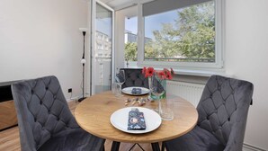 A dining area with a round table, two grey upholstered chairs, a white plate, a glass, a vase with red flowers, and a window with a view of buildings and trees.
