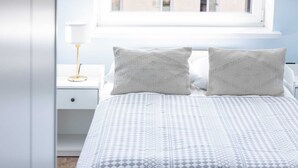 A bedroom with a patterned bedspread, two pillows, a bedside table with a lamp, and a window with a view of buildings.