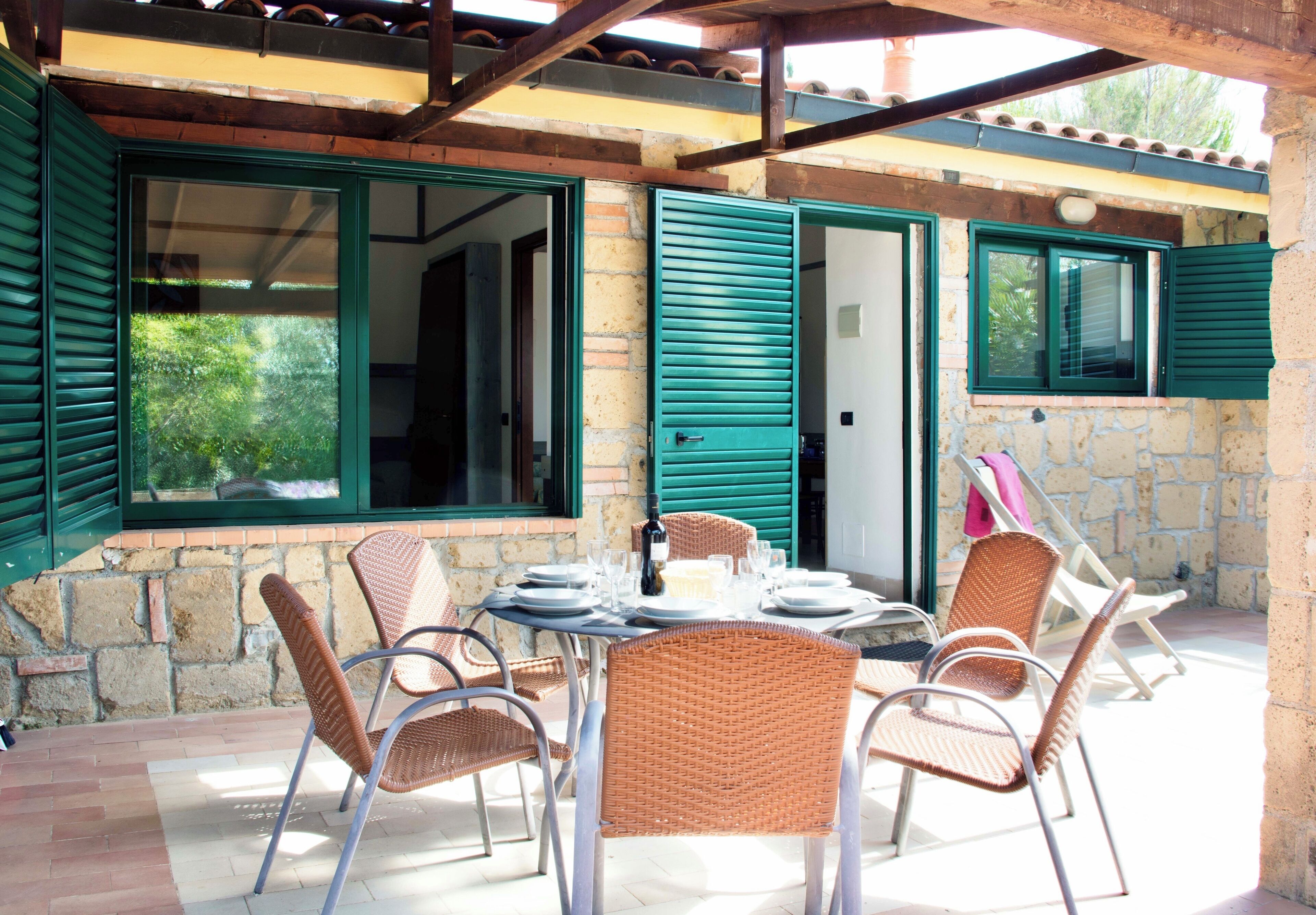A covered patio with wicker chairs and a table set for dining, featuring a stone wall and green shutters.