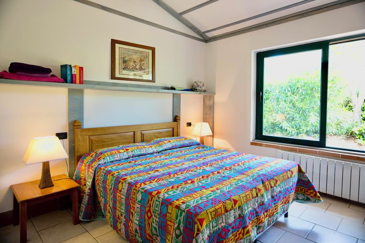 A bedroom with a bed, two bedside tables with lamps, a shelf with books and a framed picture, and a window with a view of greenery.