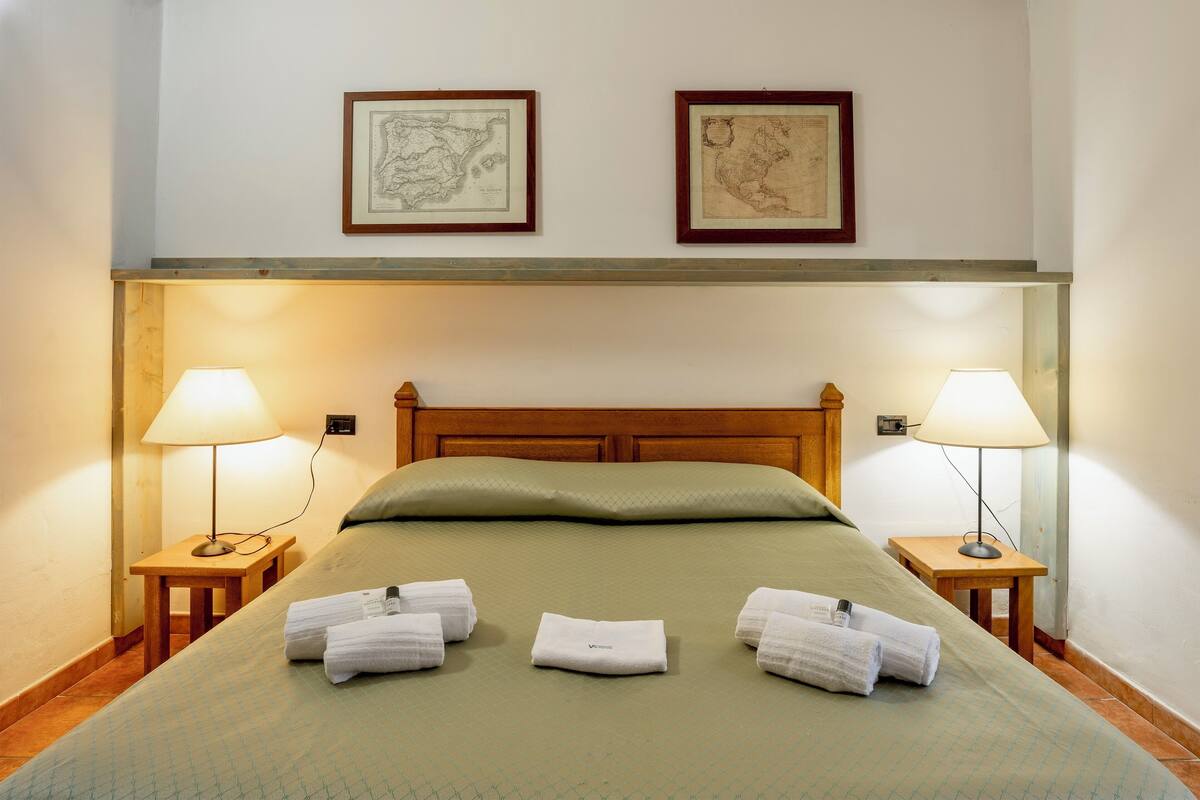 A neatly made bed with green bedding, two framed maps on the wall, and two bedside lamps.