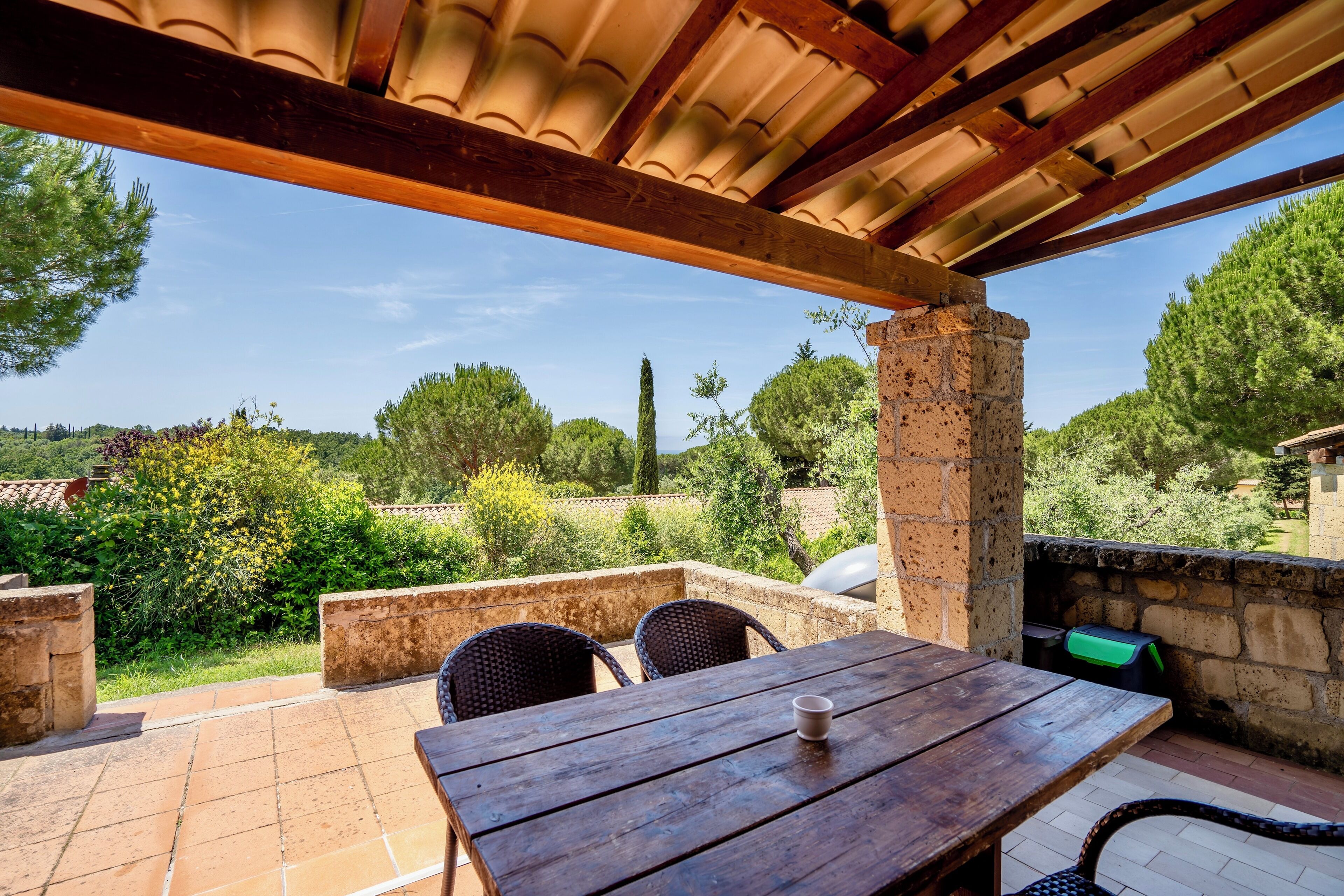 Un patio al aire libre con una mesa y sillas de madera, un muro de ladrillo y vistas a follaje y árboles.