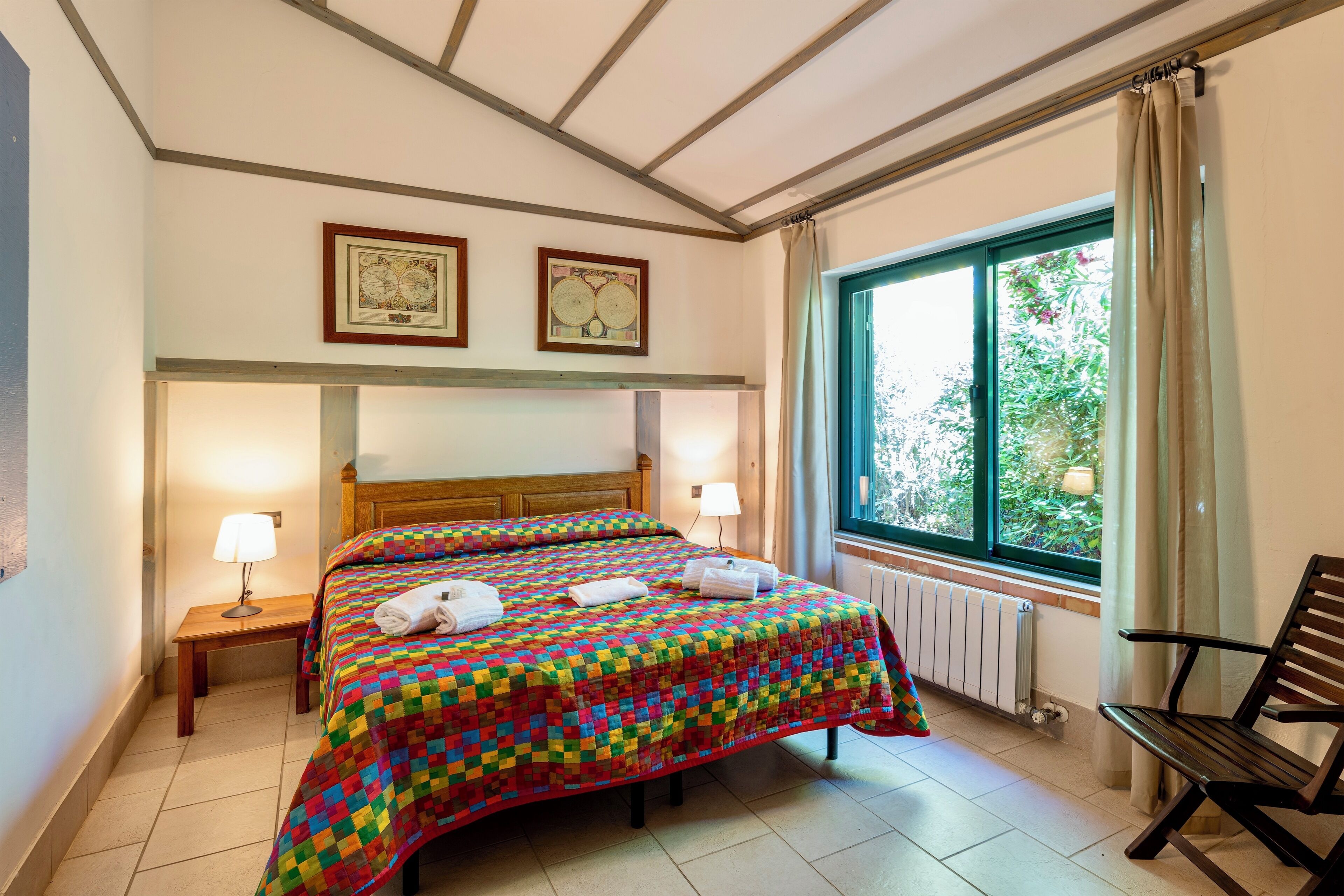 Una camera d'albergo con un letto matrimoniale, due lampade sui comodini, una testiera in legno, una finestra con tende, un termosifone e una panca.