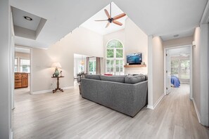 A spacious living area with a grey sofa, a ceiling fan, a TV mounted on the wall, and a large window providing natural light.