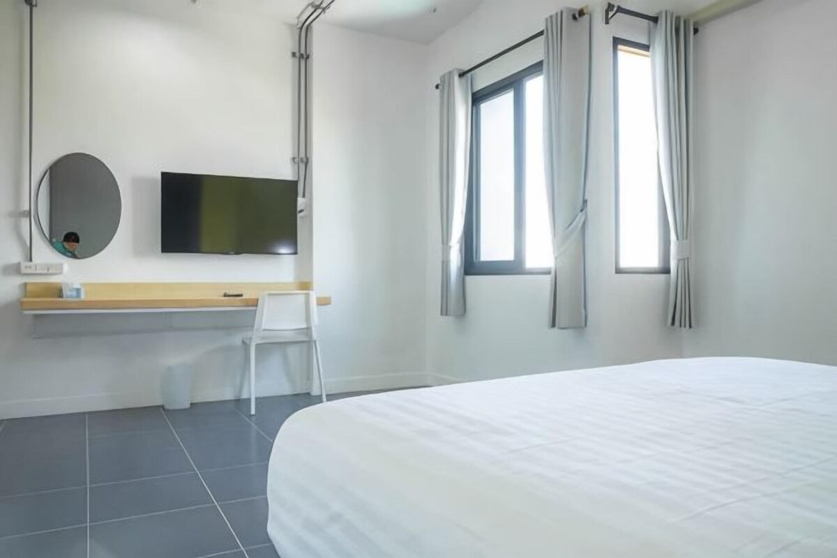 A hotel room with a bed, a desk, a chair, a TV, and two windows with curtains.