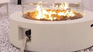 A circular outdoor fire pit with a glass enclosure, surrounded by white Adirondack chairs, set against a white fence and a sunset sky. - HotTub-Fire Pit-Outdoor Dining-EV Charger (East Wenatchee)