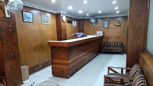 A reception area with a wooden reception desk, two chairs, a fan, and a wall-mounted clock. - Hotel Orchid (Guwahati)